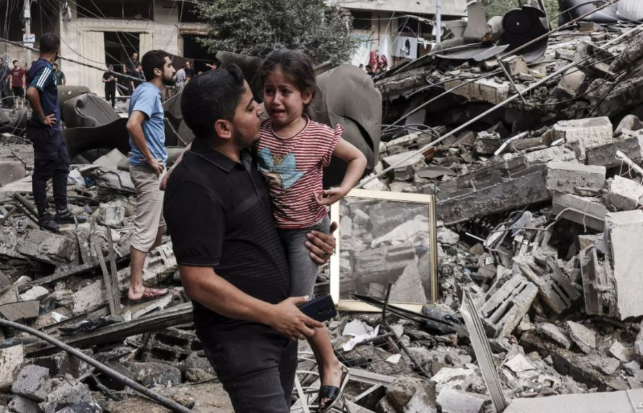 A man carries a traumatized child as he walks in front of a building destroyed in an Israeli air strike in Gaza City on October 7, 2023. (AFP)