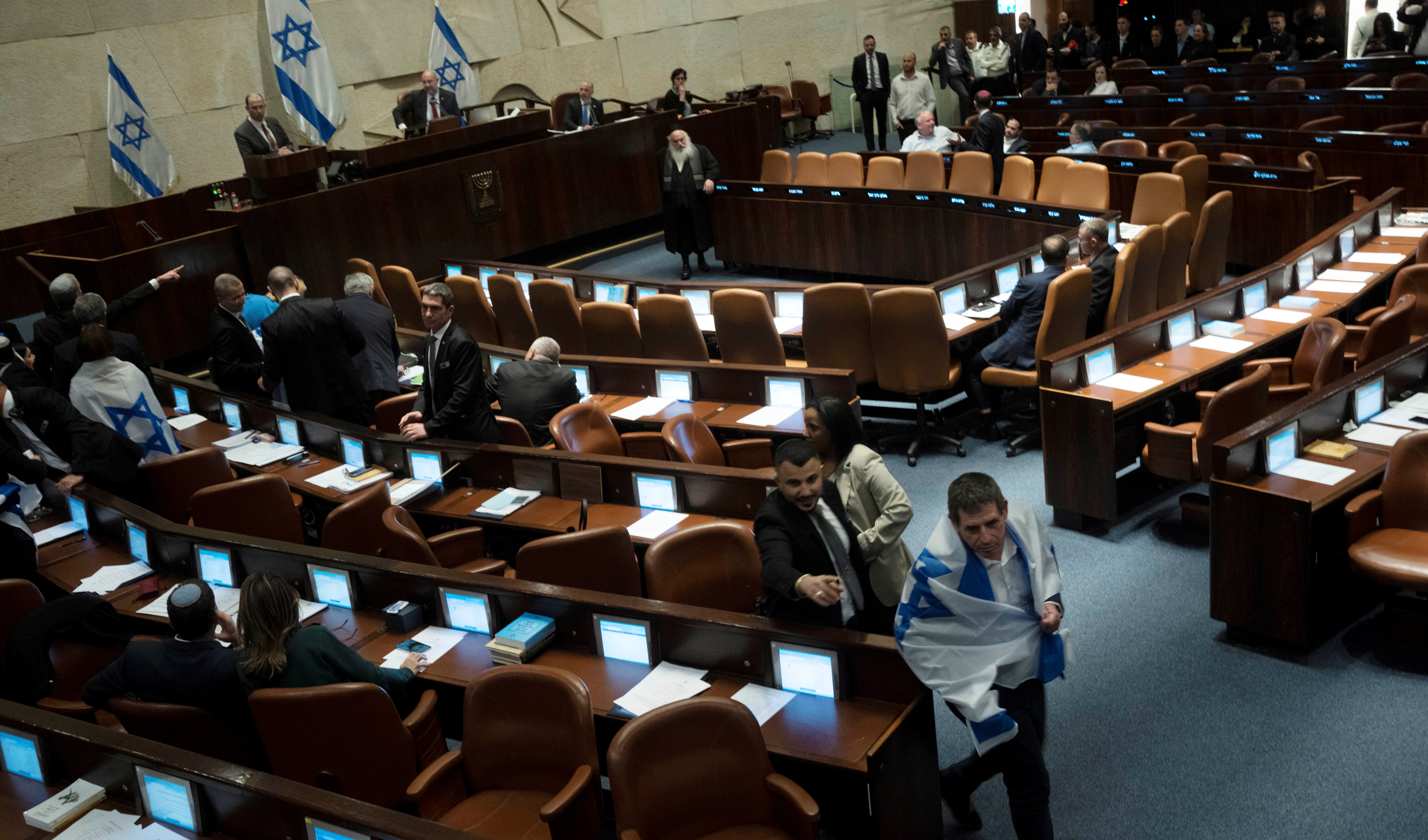 An Israeli lawmaker is pursued by security as he drapes himself in the national flag in protest, in the Knesset, against a contentious plan to overhaul the country's legal system, in Al-Quds, Monday, Feb. 20, 2023 (AP).