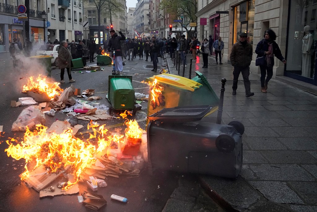 People walk by burning garbage cans during a protest, Friday, April 14, 2023 in Paris (AP Photo/Michel Euler)